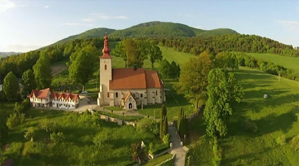 Letecký pohľad na vidiecku krajinu Gaboltov s kostolom s červenou strechou a vežou, obklopený sviežimi zelenými poliami a zvlnenými kopcami pod jasnou oblohou.