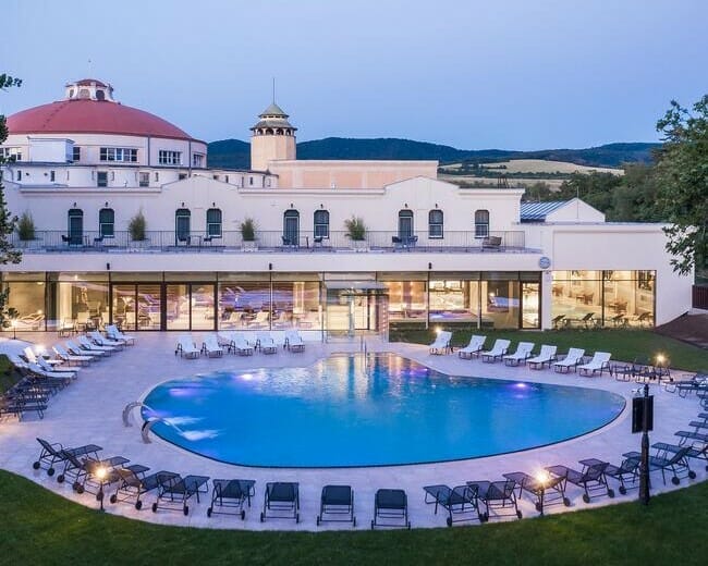Letecký pohľad na luxusný hotel za súmraku s veľkým vonkajším bazénom obklopeným ležadlami, s bujnými kopcami v pozadí pri špičkových slovenských kúpeľoch.