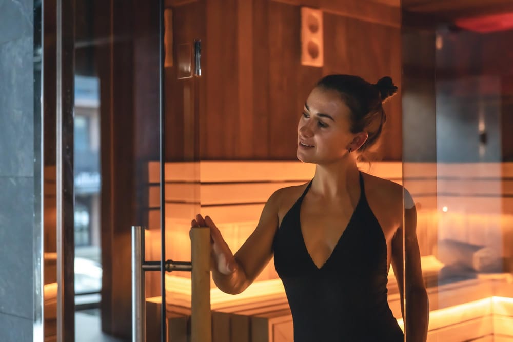 Žena v čiernych plavkách stojaca pred saunou osvetlenou infračervenou lampou.